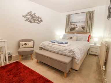 A bedroom with a bed a bench at the foot of the bed a chair with a pillow and a red rug on the floor at Angorfa in Beaumaris