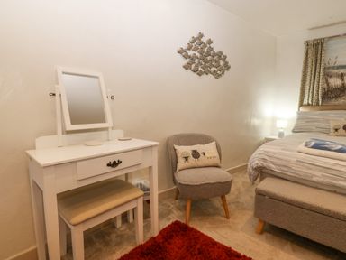 A bedroom with a white dressing table and stool a grey chair with a cushion a bed with striped bedding and a bench at Angorfa in Beaumaris