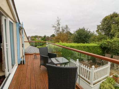 A balcony with two chairs and a small table overlooking a garden with trees and a white picket fence at Angorfa in Beaumaris