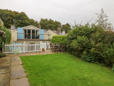 A garden with a wooden dining table and chairs a stone path green lawn and a house with blue shutters and a balcony at Angorfa in Beaumaris