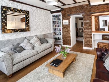 A living room with a grey sofa pillows wooden coffee table vase with flowers bookshelf stone wall and mirror at Garretts in Kingsand And Cawsand