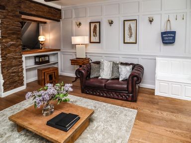 A living room with a brown leather sofa and pillows a wooden coffee table with a vase of flowers wooden floor and white paneled wall at Garretts in Kingsand And Cawsand