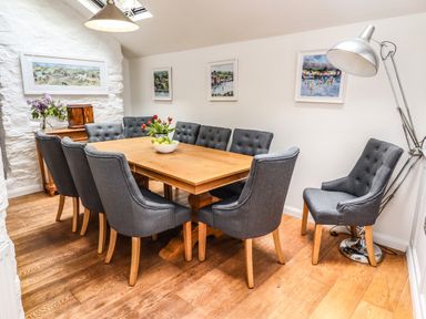 A dining room with a wooden table surrounded by gray upholstered chairs flowers and fruit on the table and framed paintings on the wall at Garretts in Kingsand And Cawsand