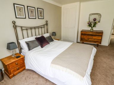 A bedroom with a bed with pillows and blanket two wooden bedside tables with lamps and a wooden dresser with flowers at Garretts in Kingsand And Cawsand