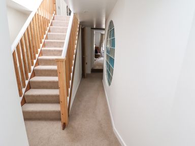 A narrow hallway with beige carpet and wooden staircase next to a circular glass block window at Garretts Kingsand And Cawsand