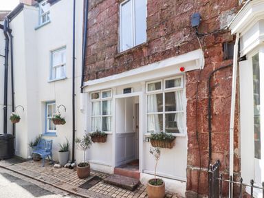 The entrance of a house with white framed windows and door with plants in pots and hanging baskets outside Garretts Kingsand And Cawsand
