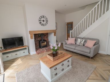 A living room with a gray sofa and pink cushions a TV on a stand a wood-burning stove and a coffee table with flowers at The Miners Cottage in Scremerston near Berwick-Upon-Tweed