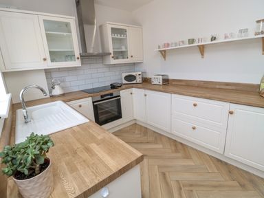 A kitchen with white cabinets wooden countertops a sink stove microwave toaster a plant and mugs on a shelf at The Miners Cottage in Scremerston near Berwick-Upon-Tweed