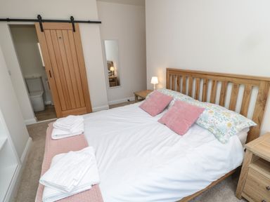 A bedroom with a wooden bed frame, white bedding, pink pillows, bedside tables, a lamp, and a wooden sliding door at The Miners Cottage in Scremerston near Berwick-Upon-Tweed