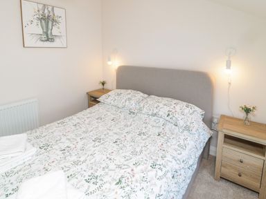 A bedroom with a bed with floral bedding two wooden bedside tables with lamps and flowers and a painting on the wall at The Miners Cottage in Scremerston near Berwick-Upon-Tweed