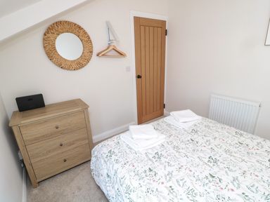 A bedroom with a bed covered in floral bedding and folded towels a wooden chest of drawers a round woven mirror a wooden door and a radiator at The Miners Cottage in Scremerston near Berwick-Upon-Tweed