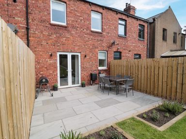 A backyard patio with a table and six chairs a small barbecue potted plants flower beds and a wooden fence at The Miners Cottage in Scremerston near Berwick-Upon-Tweed