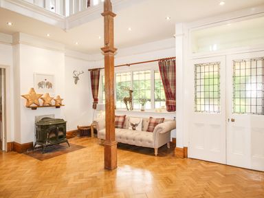 A sitting area with a sofa and cushions next to a window with curtains and a wood stove in a room with wooden flooring at Deer Leap House in Moreton