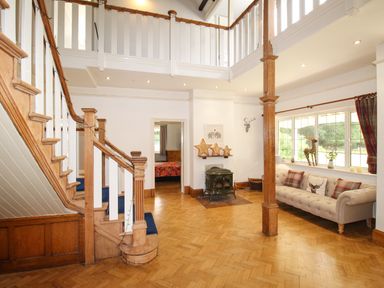 An entrance hall with wooden staircase a beige sofa with cushions a wood burning stove and large windows at Deer Leap House in Moreton