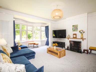 A living room with a blue sofa and bay windows a wooden coffee table flowers on the table a fireplace with a mantel and a wall-mounted television at Deer Leap House in Moreton