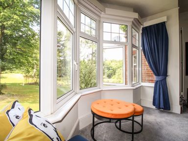 A corner with large bay windows showing a garden outside two round orange stools and a blue curtain at Deer Leap House in Moreton