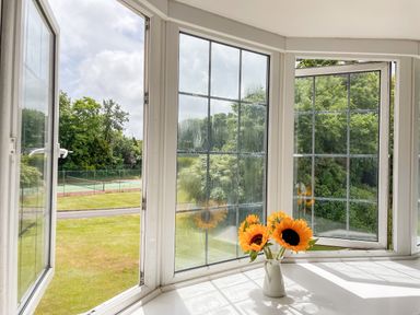 A white vase with sunflowers on a windowsill with open windows overlooking a green lawn and tennis court at Deer Leap House in Moreton