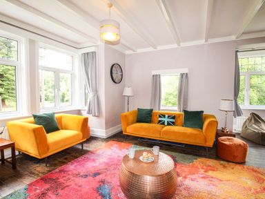 A living room with two yellow sofas green cushions a round wooden coffee table colorful rug and large windows at Deer Leap House Moreton