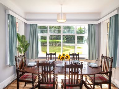 A dining room with six chairs around a wooden table set with plates and glasses and sunflowers in a vase at Deer Leap House in Moreton
