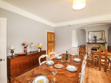 A dining room with a wooden table set with dishes glasses and cutlery beside a wooden sideboard with lamps bowls of fruit and flowers at Deer Leap House in Moreton