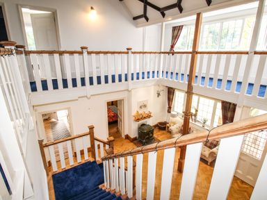 A staircase with blue carpet leading to an upper hallway with white railings and blue carpet overlooking a living room with armchairs and large windows at Deer Leap House in Moreton