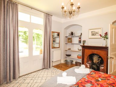 A bedroom with a bed covered in a red and gray blanket rolled white towels a fireplace shelves with decorative items and glass doors with curtains at Deer Leap House in Moreton