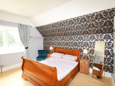 A bedroom with a wooden bed and matching bedside tables a blue upholstered chair a tall mirror and patterned wallpaper at Deer Leap House in Moreton