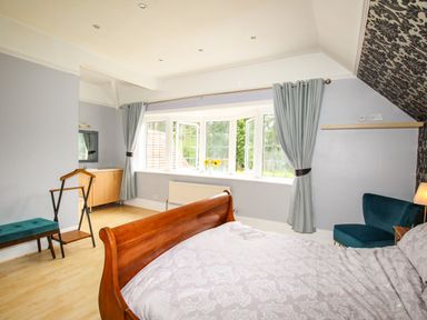 A bedroom with a wooden bed frame blue chair and bay window with curtains at Deer Leap House in Moreton