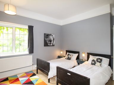 A bedroom with two single beds with white bedding and black headboards a colorful geometric rug on the floor a window with dark curtains a black and white lion picture on the wall and two bedside tables with lamps at Deer Leap House in Moreton