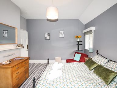 A bedroom with a bed with geometric patterned bedding a wooden dresser a red chair with a blue cushion and a window with blinds at Deer Leap House in Moreton