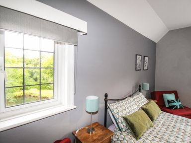A bedroom with a patterned bed, green pillows, a wooden bedside table with a lamp, a red chair, and a window showing greenery at Deer Leap House in Moreton