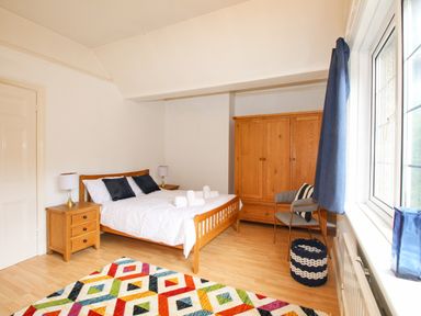 A bedroom with a wooden bed a colorful rug wooden wardrobe and a window with blue curtains at Deer Leap House in Moreton