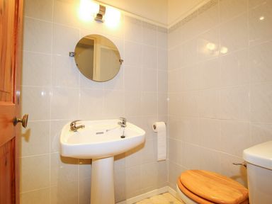 A bathroom with a pedestal sink a round mirror a toilet with a wooden seat and a toilet paper holder at Deer Leap House in Moreton