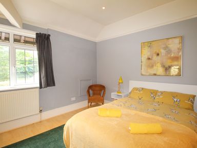 A bedroom with a double bed with yellow bedding a wicker chair a bedside table with a yellow lamp and a window with curtains at Deer Leap House in Moreton