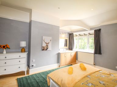 A bedroom with a bed featuring yellow bedding and towels a white dresser with a lamp and flowers a wall painting a green rug and a wooden vanity under a window at Deer Leap House in Moreton