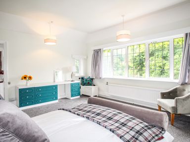 A bedroom with a bed a dresser with teal drawers a chair and large windows at Deer Leap House in Moreton