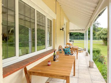 A covered outdoor patio with wooden tables and chairs alongside a grassy garden at Deer Leap House in Moreton