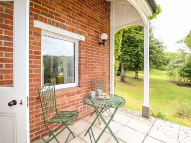 A porch with a green metal table and two chairs next to a brick wall and a door overlooking a grassy garden at Deer Leap House in Moreton