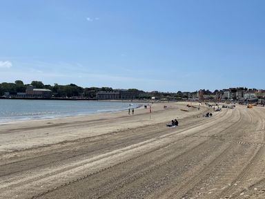 A sandy beach with people sitting and walking near the water and buildings in the background at Deer Leap House in Moreton