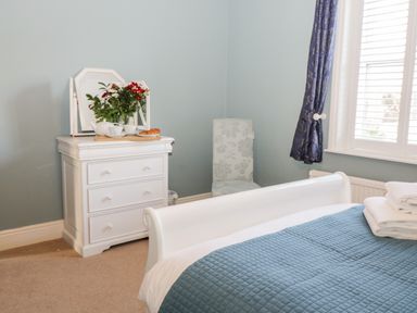 A bedroom with a white dresser holding flowers and tea set next to a window with blue curtains and a bed with folded towels at Hazeldene in Sleights