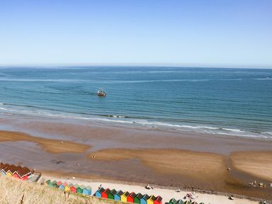 A beach with colorful beach huts along a promenade and a ship sailing on the sea at Hazeldene in Sleights