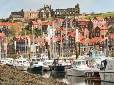 A harbor with multiple small boats docked in front of red-roofed houses and an old stone abbey on a hill