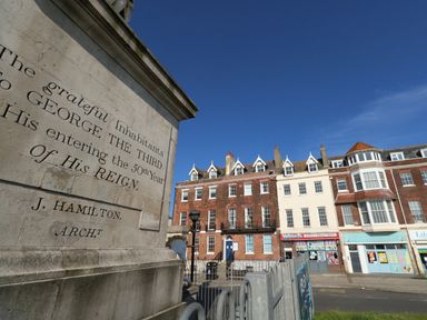 A street view with a stone monument engraved with text and row of mixed brick and white buildings with shops below a clear blue sky at The Kings Statue in Weymouth