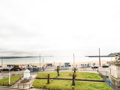 A beach view with a road, green grass, palm trees, a statue, and the sea at The Kings Statue in Weymouth