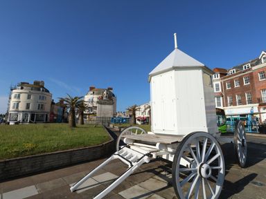 A white wooden cart with large wheels on a paved area near a grassy roundabout with palm trees and a statue at The Kings Statue in Weymouth