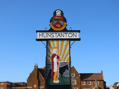 A sign with the word Hunstanton and a colorful image of a crowned figure with a dog and rays of light in front of buildings at Maples in Hunstanton