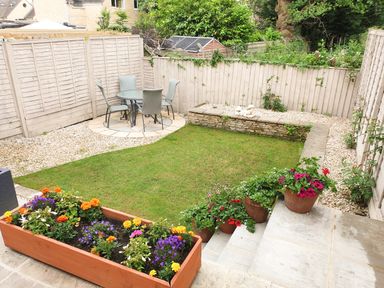 A garden with a small lawn, flower pots on stone steps, a round table and four chairs on a gravel and paved area at The Seventeen in Bourton-On-The-Water