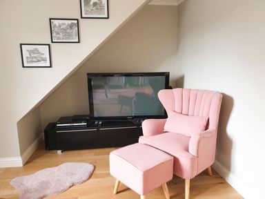 A pink armchair with matching footstool near a black TV stand with a television and framed pictures on the wall at The Seventeen in Bourton-On-The-Water