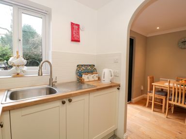 A kitchen sink with a window above and a kettle on the counter next to a dining area with a wooden table and chairs at The Seventeen in Bourton-On-The-Water