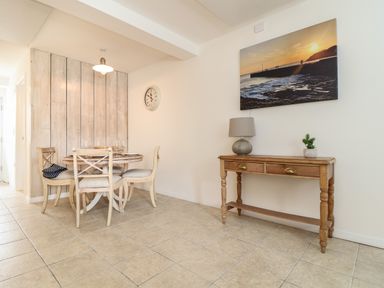 A dining area with a round table, four chairs, a wall clock, a wooden console table with a lamp and a plant, and a sunset seaside picture at The Old Post Office in Porthleven